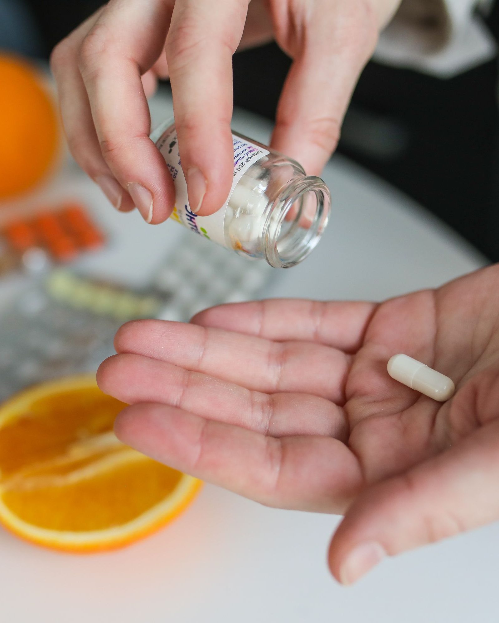 Vue de dessus : une main tient une plaquette de capsules orange, un verre d’eau posé sur une table blanche, un plaid beige